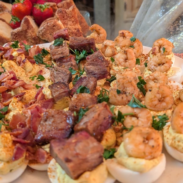 Deviled eggs topped with shrimp, steak bites, and bacon, garnished with parsley and paprika.