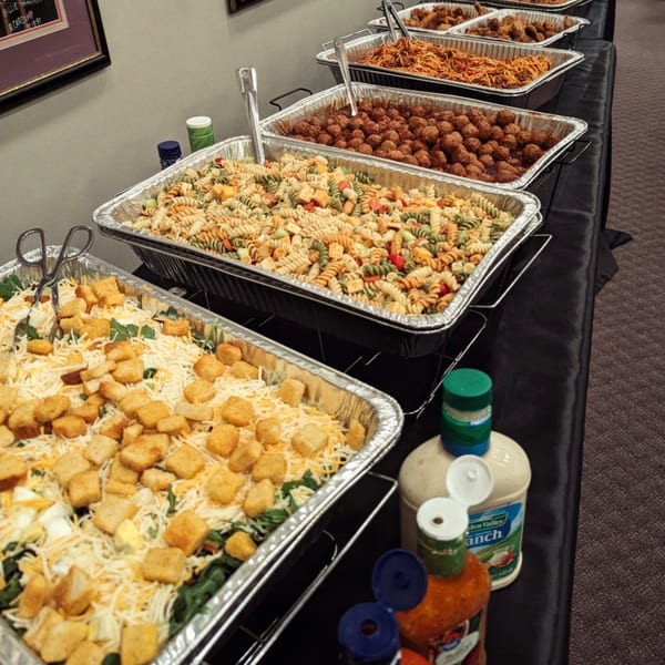 A buffet line with trays of salad, pasta salad, meatballs, spaghetti, and chicken wings.