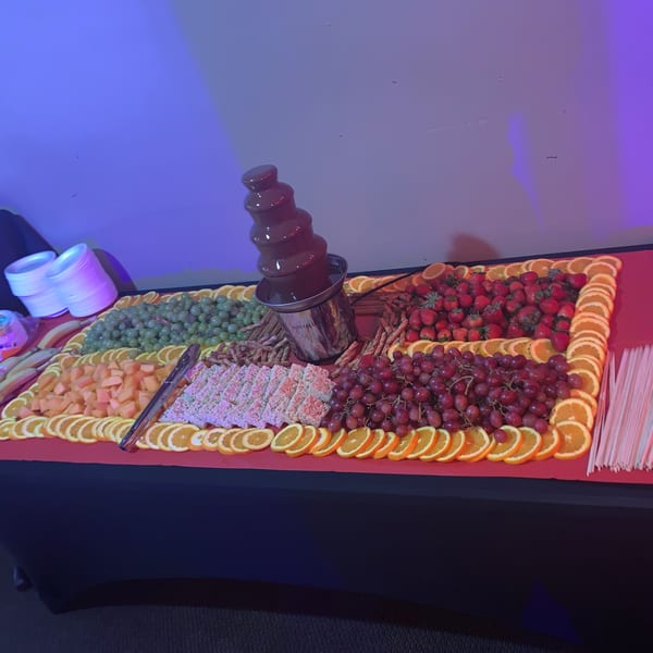 A chocolate fountain surrounded by fresh fruit, rice crispy treats, and pretzel sticks on a red tablecloth.