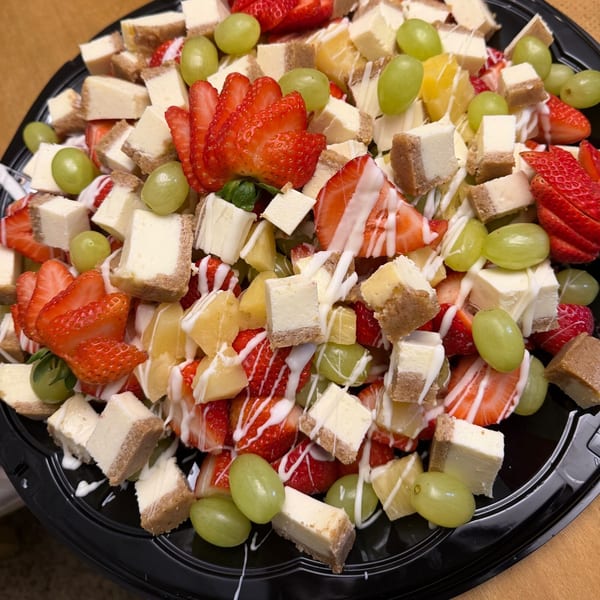 A platter of cheesecake bites, strawberries, grapes, and pineapple, drizzled with white icing.