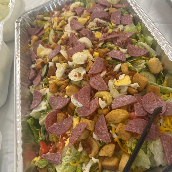 A large catering tray filled with a vibrant salad, topped with salami, eggs, croutons, and bacon bits.